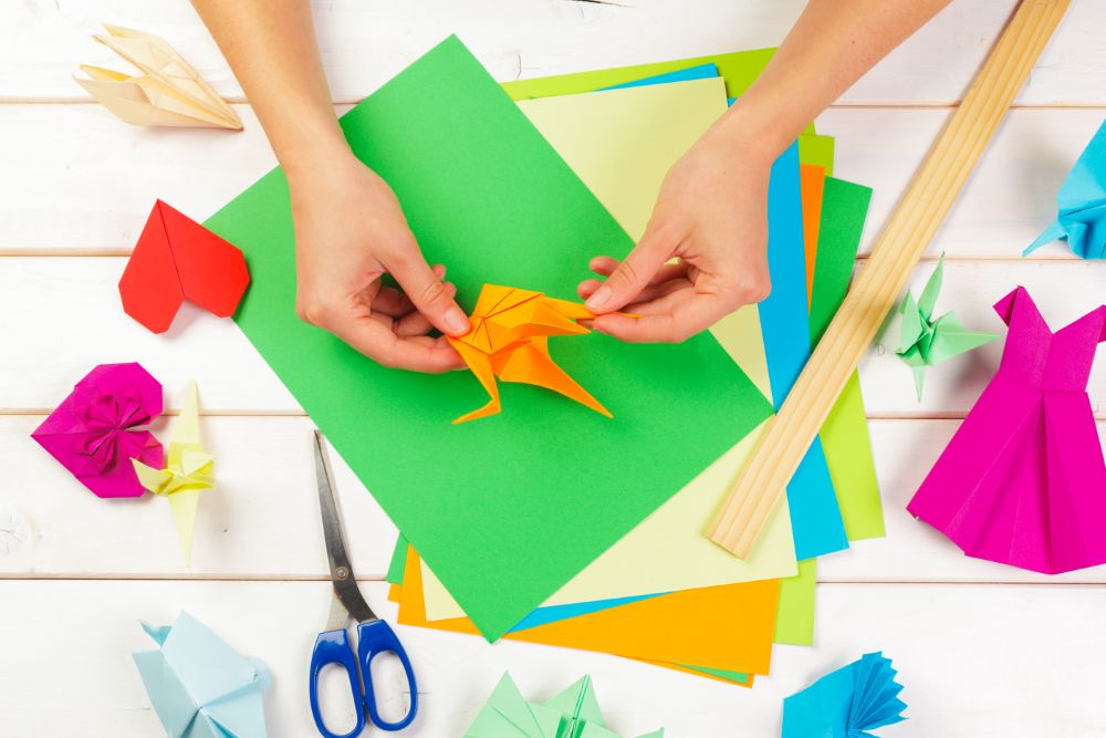 Δημιουργώντας τέχνη με papercraft Χέρια διπλώνουν ένα πορτοκαλί χαρτί σε ένα χρωματιστό τραπέζι εργασίας.